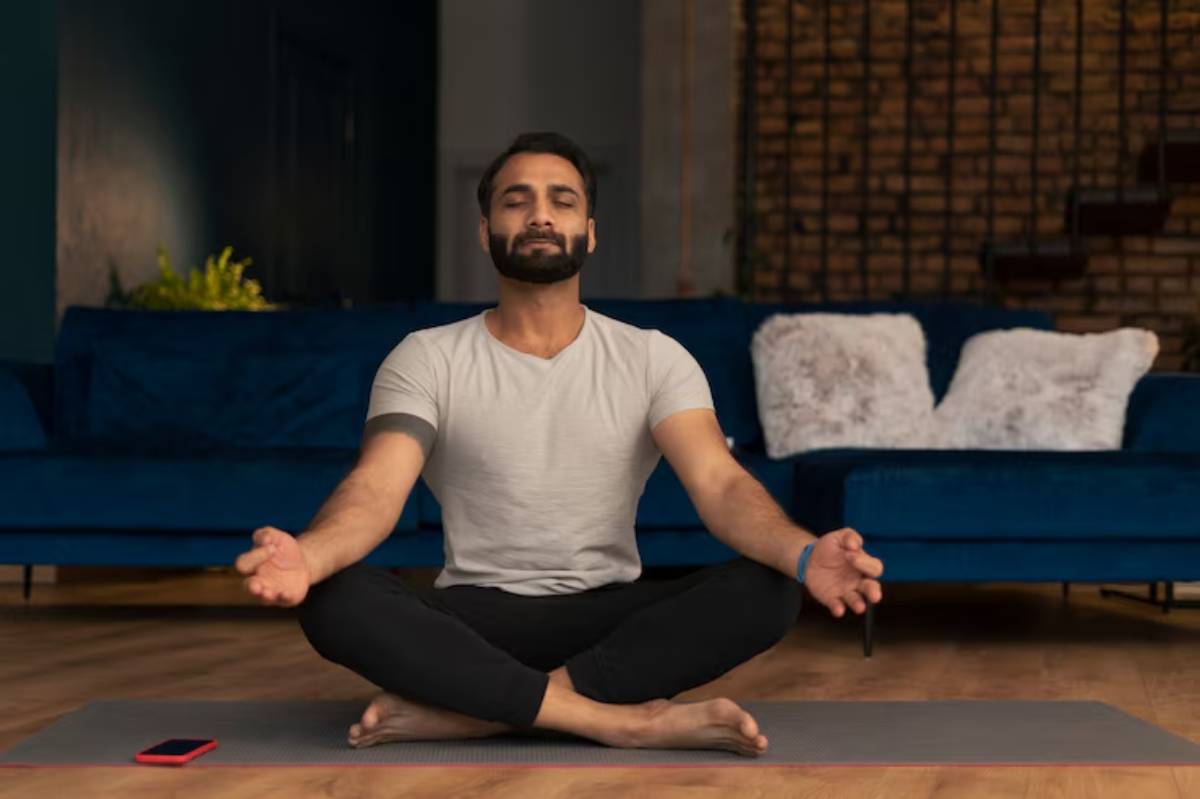 A person sitting cross-legged on a yoga mat, meditating in a cozy living room with a blue couch and decorative pillows.