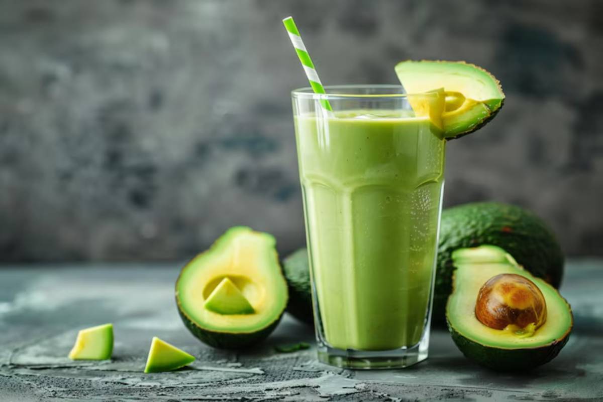 A tall glass of creamy green avocado smoothie with a striped straw, surrounded by fresh avocado halves and slices on a textured surface.
