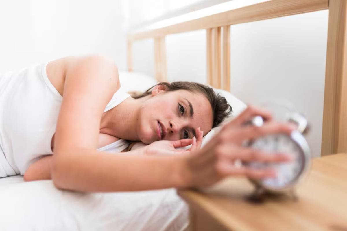 A person lies in bed, reaching for an alarm clock and try to snooze it on the nightstand, surrounded by a bright and softly lit room.