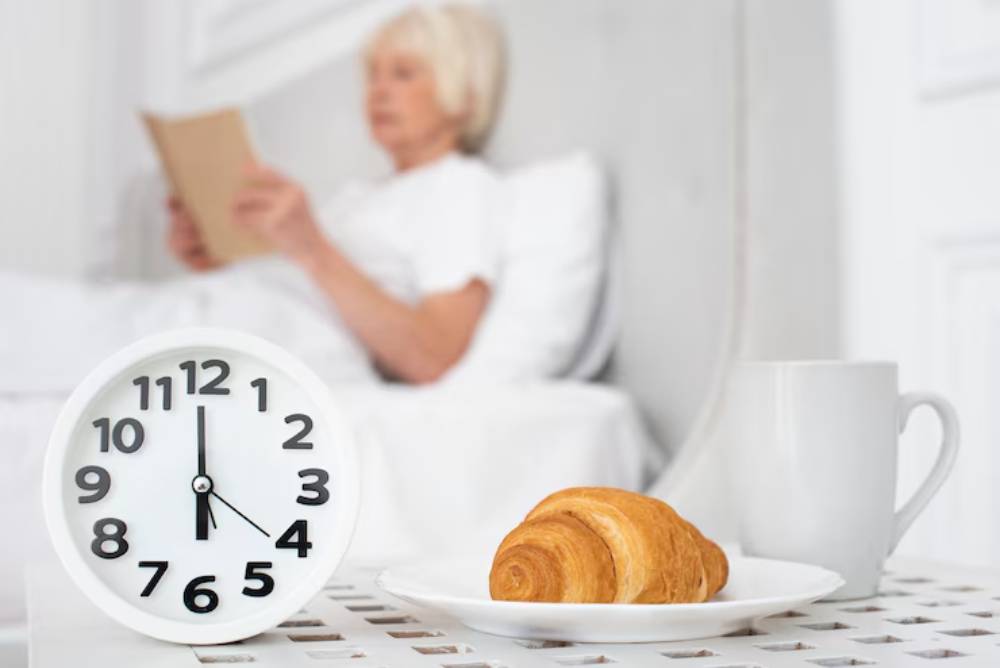 A white clock shows the time nearing 6:00, beside a plate with a croissant and a cup of coffee on a bedside table.
