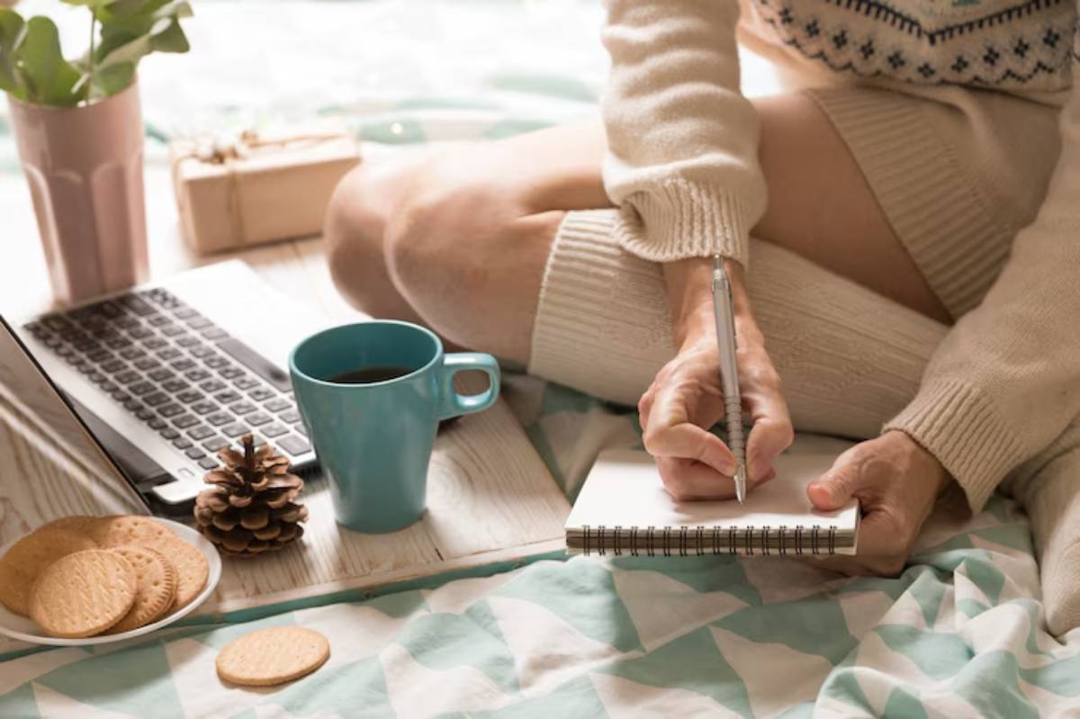 A person in a cozy sweater sits on a bed, writing in a notebook with a laptop and snacks nearby, surrounded by a serene atmosphere.