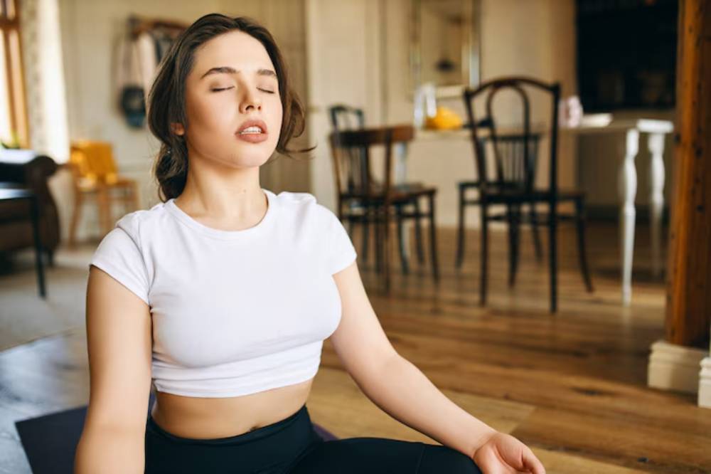 A woman sits cross-legged on a yoga mat in a cozy, well-lit room with wooden floors, surrounded by chairs and a dining table.