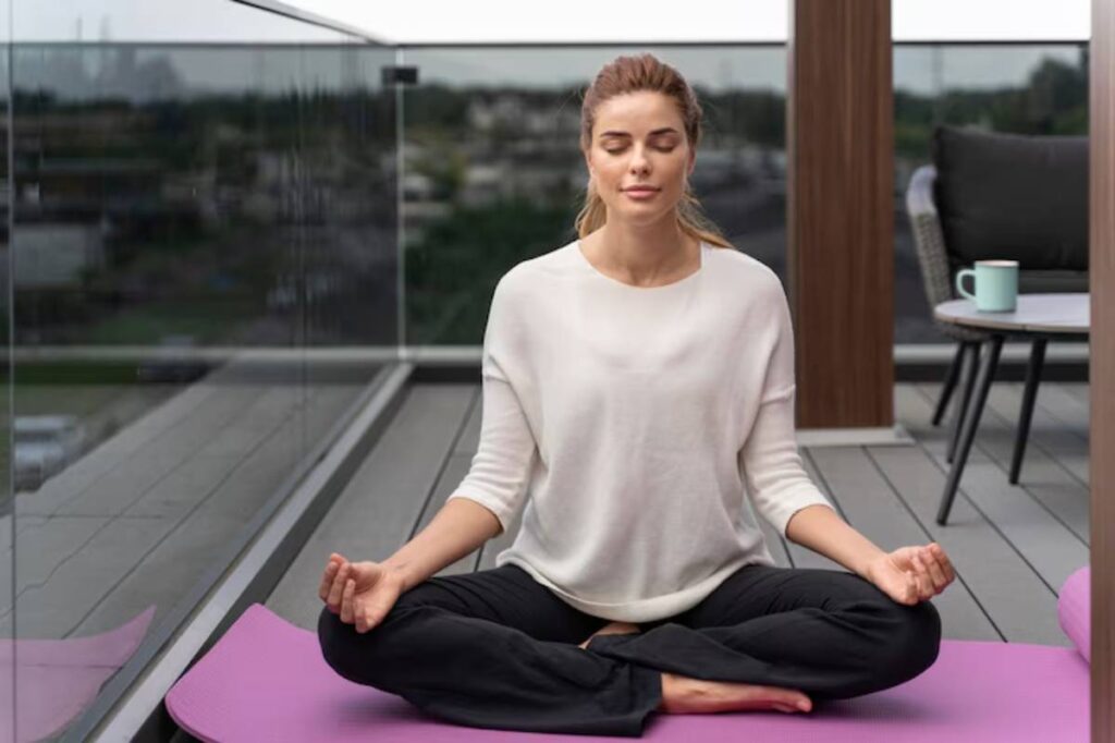 A person meditates cross-legged on a purple yoga mat on a balcony, surrounded by greenery and modern furniture.