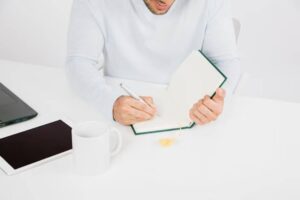 A man in a light sweater writes in a notebook while seated at a white desk with a laptop and a mug nearby.