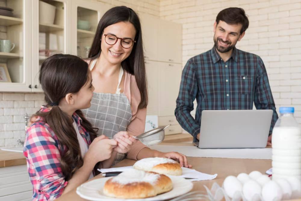 A woman in an apron interacts with a girl in a checkered shirt, while a man works on a laptop nearby in a bright kitchen.
