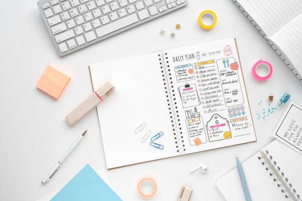 A flat lay of a planner with a colorful daily plan, surrounded by stationery, a keyboard, and a notebook on a clean workspace.