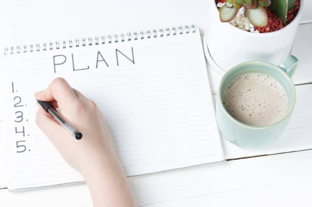 A hand writes a numbered list titled 'PLAN' in a notebook, next to a cup of coffee and a small potted plant.