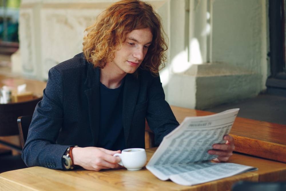 A person with curly hair sits at a wooden table, sipping coffee while reading a newspaper in a cozy café setting.