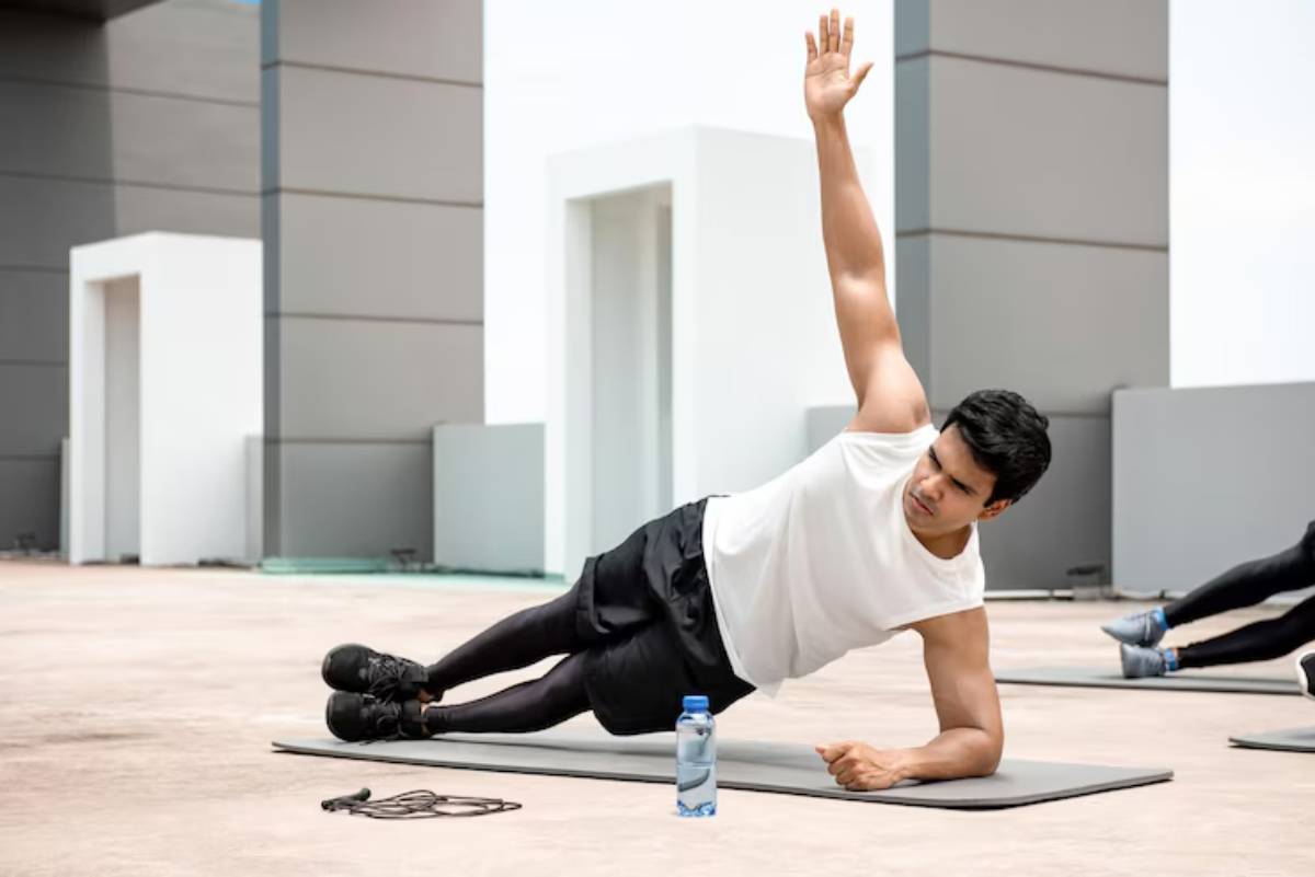 A person performs a side plank exercise on a mat, raising one arm, with a water bottle nearby and workout equipment on the ground.