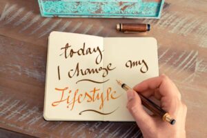A hand writes 'today I change my lifestyle' in a notebook, alongside a fountain pen and decorative box on a wooden table.