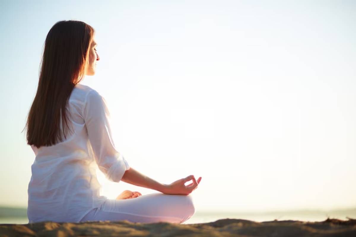 A woman in a white outfit sits cross-legged on the beach, meditating against a backdrop of soft sunlight and a calm horizon.
