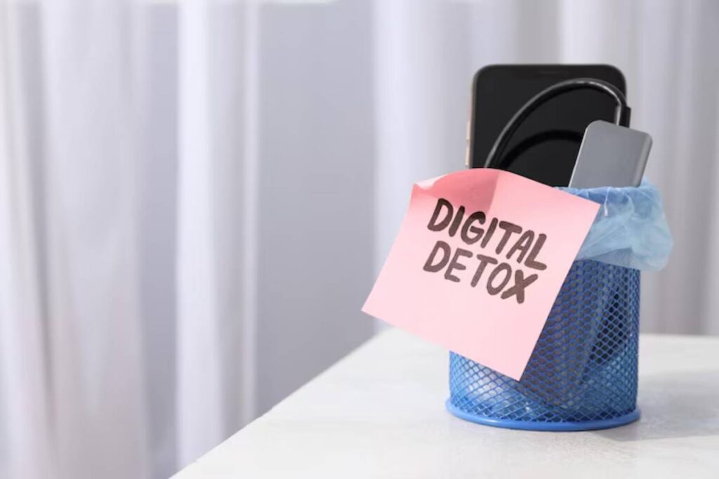 A blue mesh trash can holds a smartphone and charger, topped with a pink note reading 'DIGITAL DETOX.'