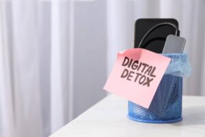 A blue mesh trash can holds a smartphone and charger, topped with a pink note reading 'DIGITAL DETOX.'