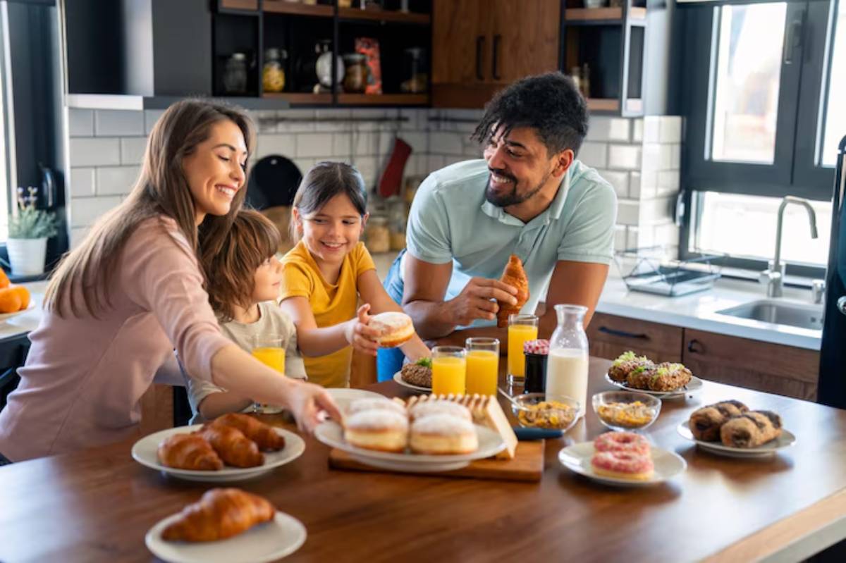 A family enjoys a breakfast spread on a wooden table, featuring croissants, donuts, juice, and other pastries in a bright kitchen.