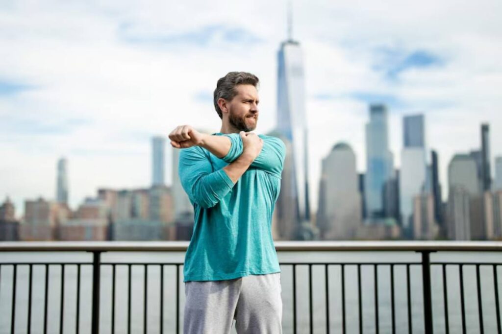 A man stretches by a waterfront with a city skyline and skyscrapers in the background, wearing a turquoise long-sleeve shirt and gray pants.