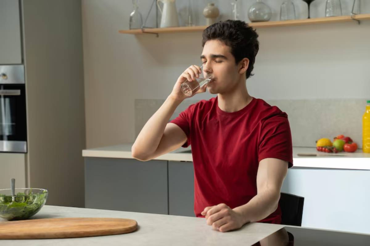 A person in a red shirt sits at a kitchen table, holding a glass of water, with fresh fruits and a bowl of salad nearby.