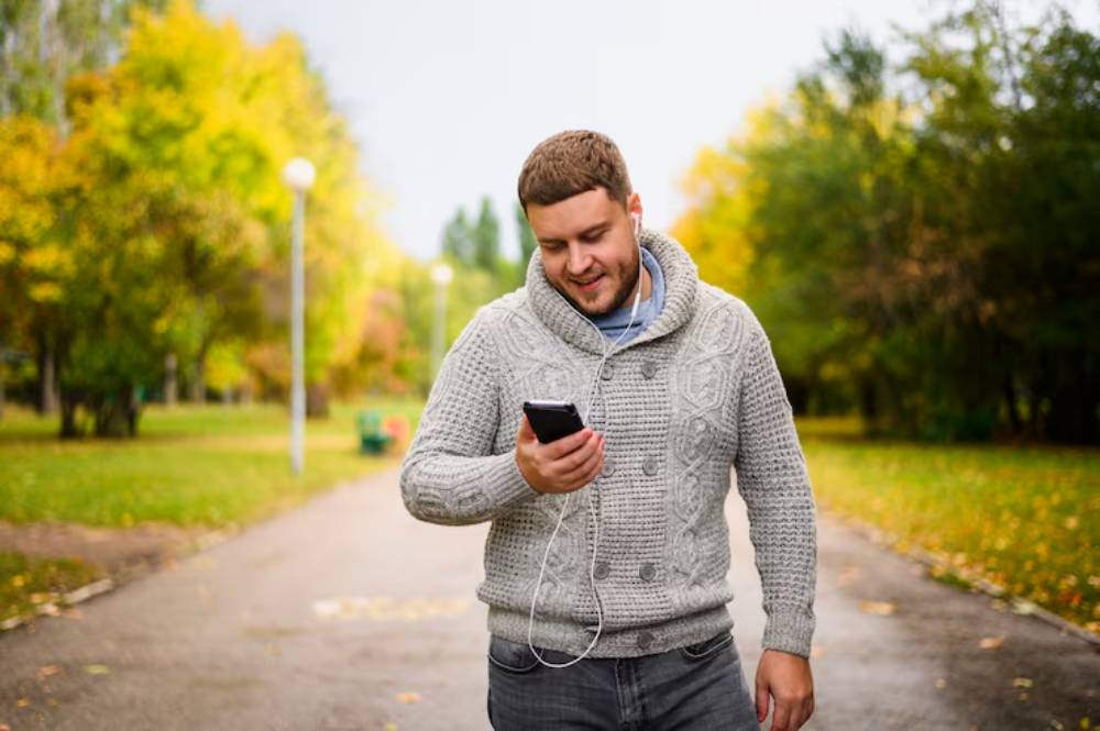 A person in a cozy sweater listens audiobook on their phone while walking down a scenic park path lined with colorful autumn trees.