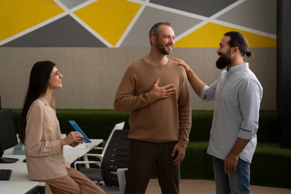A woman sits at a desk with a tablet while two men stand nearby, one with a hand on his chest and the other gratitudes.