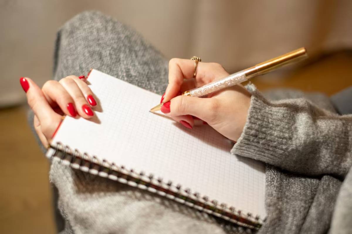 A hand with red nails holds a gold pen, writing in a blank notebook while resting on a cozy gray blanket.