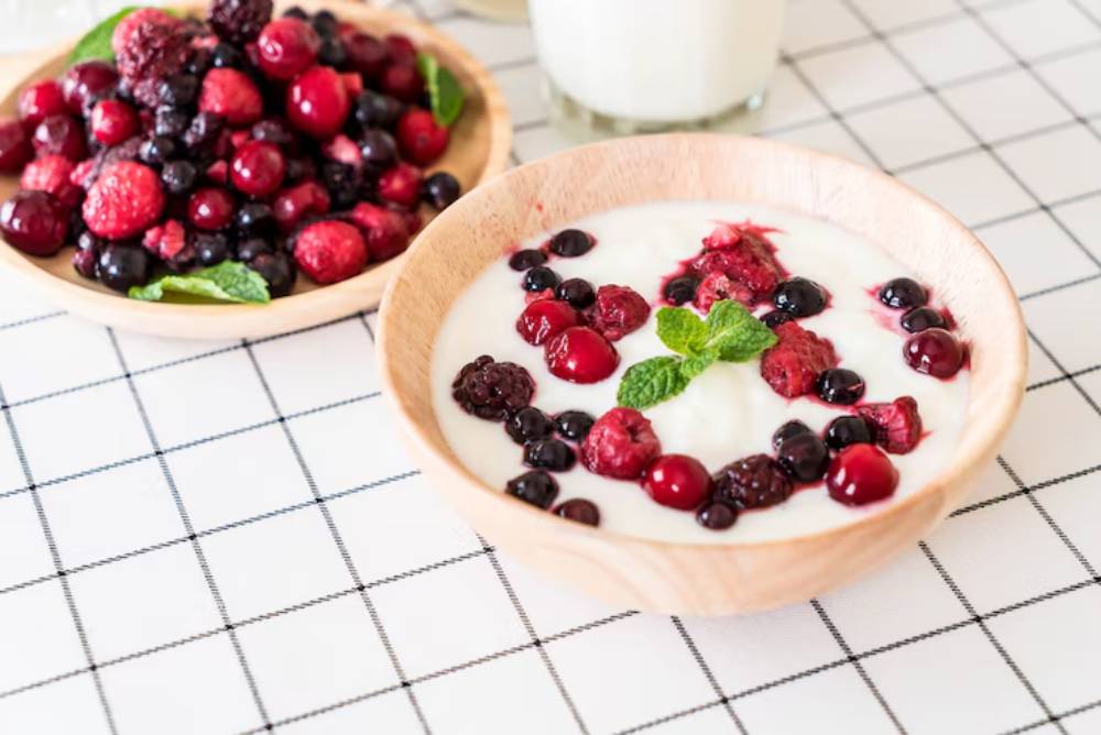A wooden bowl filled with creamy yogurt topped with mixed berries and a mint leaf, alongside a bowl of fresh berries on a checkered tablecloth.