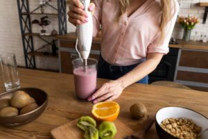 A woman blends a pink smoothie in a glass, surrounded by fresh fruits and a bowl of granola on a wooden kitchen table.