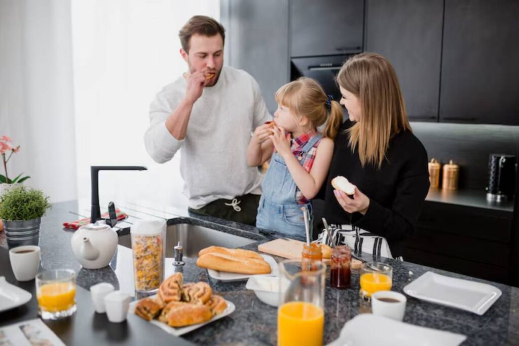 A family enjoys breakfast together in a modern kitchen. The table is filled with pastries, cereals, and beverages.