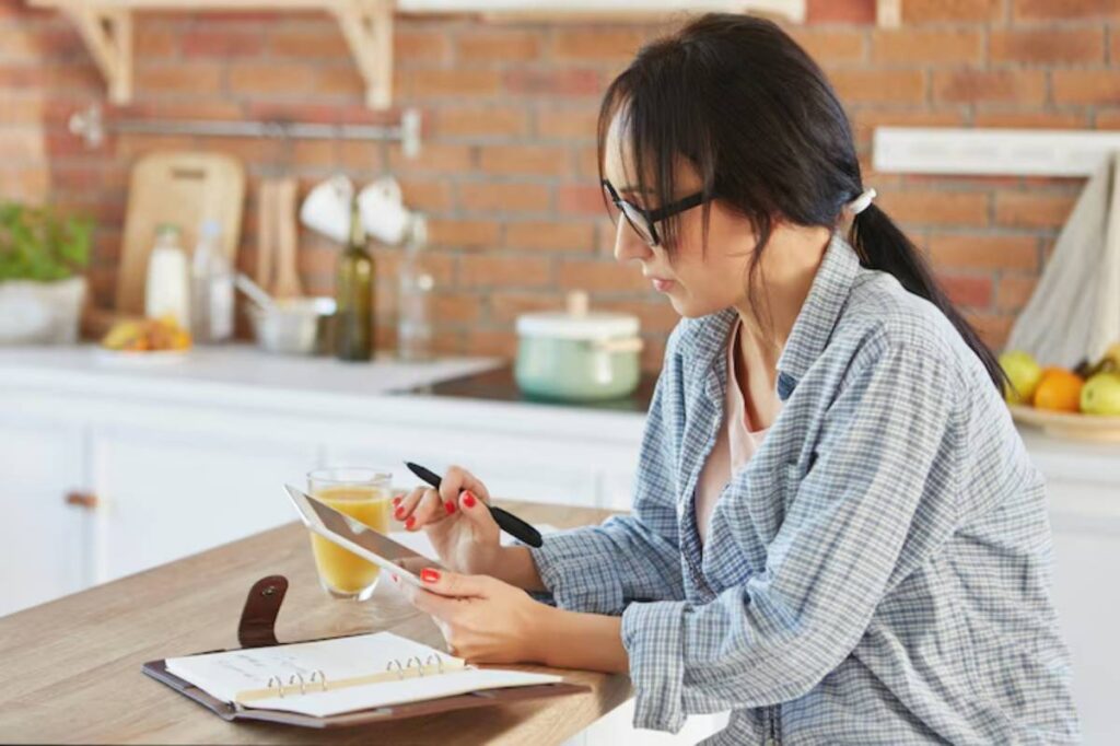 A person in a checkered shirt sits at a kitchen island, holding a tablet and a pen, with a glass of juice and a notepad nearby.