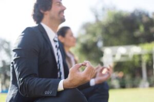 Two individuals in business attire practicing meditation outdoors, seated on grass with hands in a meditative gesture.