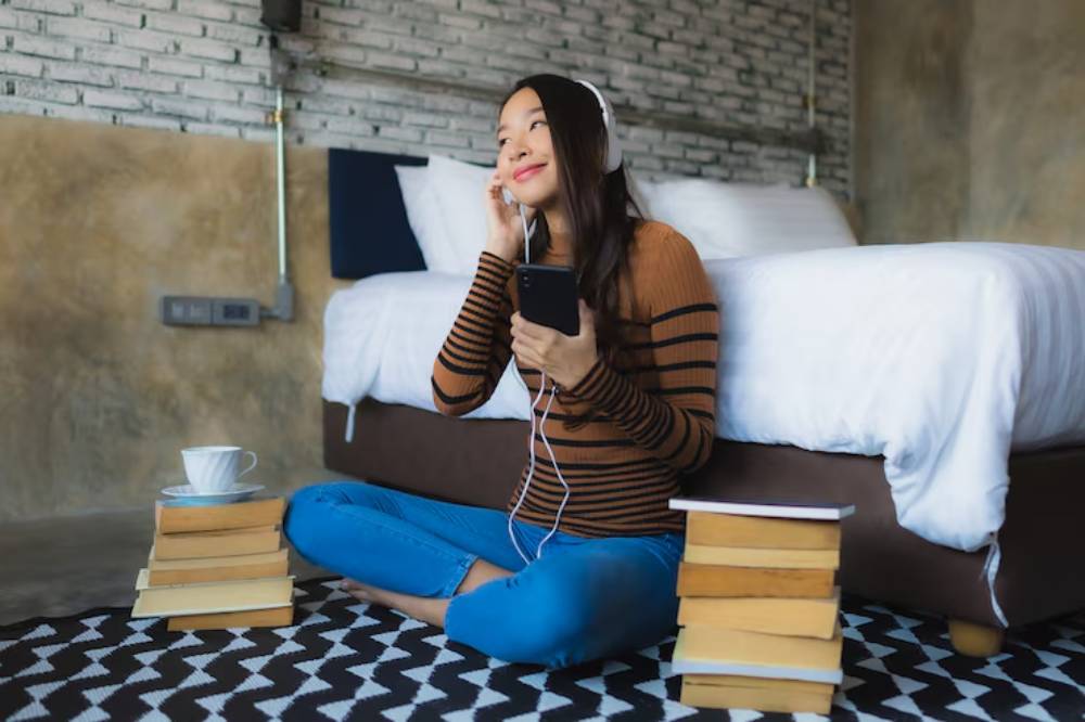 A person sits cross-legged on a rug, wearing headphones, holding a phone, with books stacked nearby and a cup of tea beside them.