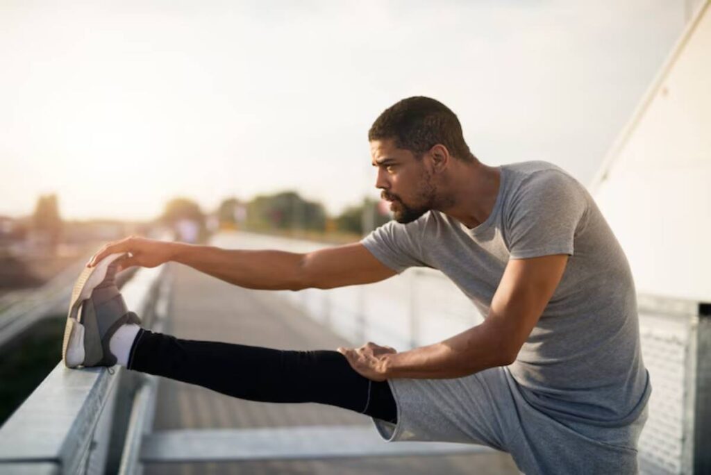 A man in a gray athletic outfit stretches his leg on a railing during sunset, with a serene landscape in the background.