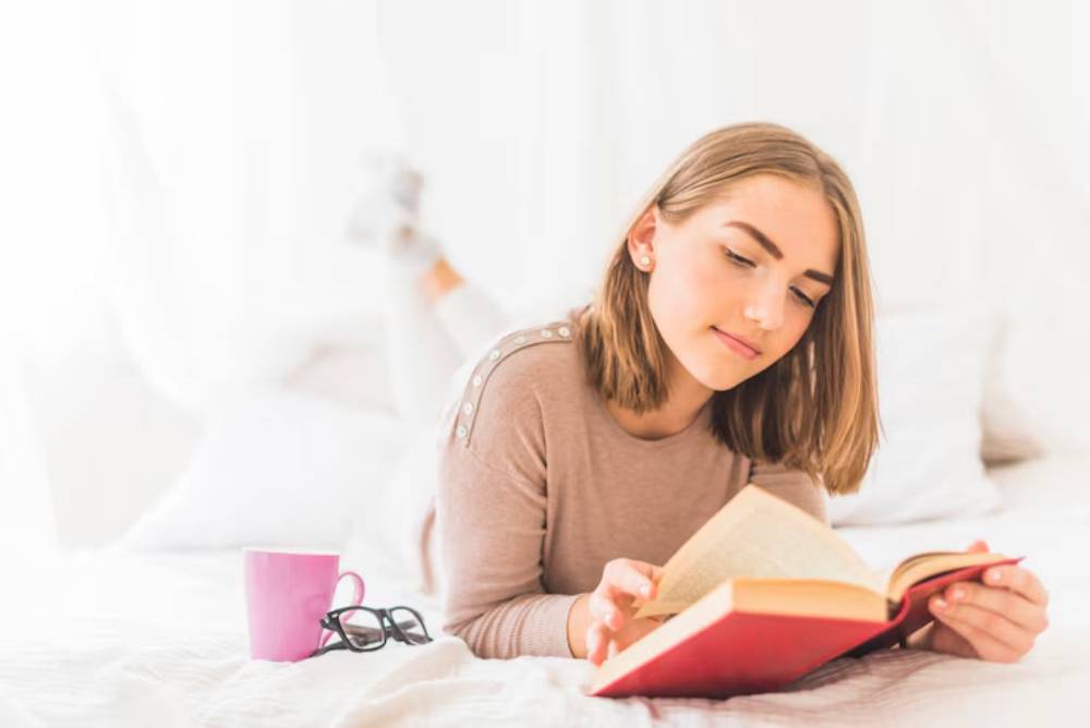 A person lies on a bed, reading a book with a pink mug beside them, surrounded by soft white bedding and natural light.