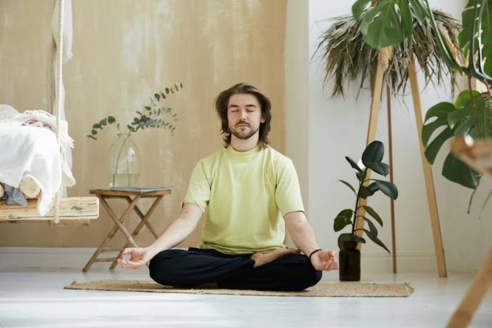 A person sits cross-legged in a peaceful room, practicing meditation, surrounded by greenery and natural decor.