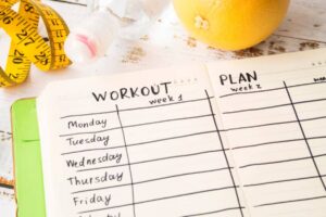 A fitness planner open to workout and meal plan sections, with a measuring tape, water bottle, and a grapefruit beside it.