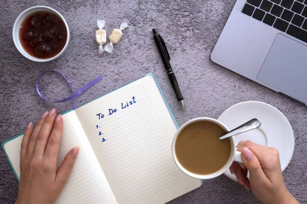 A person's hand holds a cup of coffee above an open notebook titled 'To Do List,' surrounded by treats and a laptop.