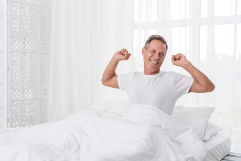 A person stretches in bed, surrounded by fluffy white blankets and natural light streaming through sheer curtains.