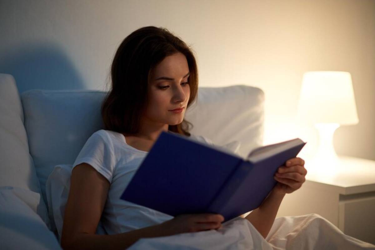 A person sits in bed reading a purple book, illuminated by a nearby lamp, surrounded by soft white bedding and a calm atmosphere.