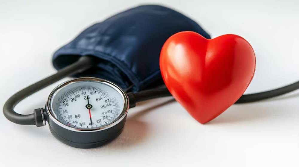 A blood pressure monitor beside a shiny red heart symbol, representing cardiovascular health and wellness.