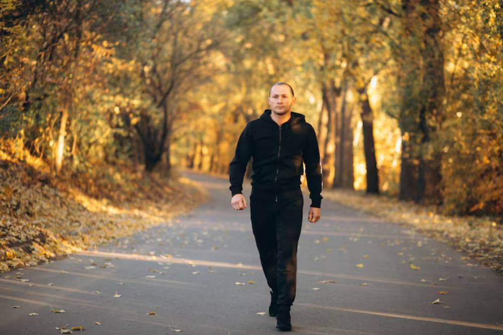 A person in a black outfit walks leisurely down a tree-lined path adorned with autumn leaves, bathed in warm sunlight.