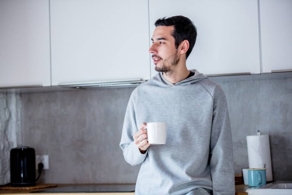 A person in a gray hoodie sits on a kitchen counter, holding a white mug, with a modern kitchen backdrop.