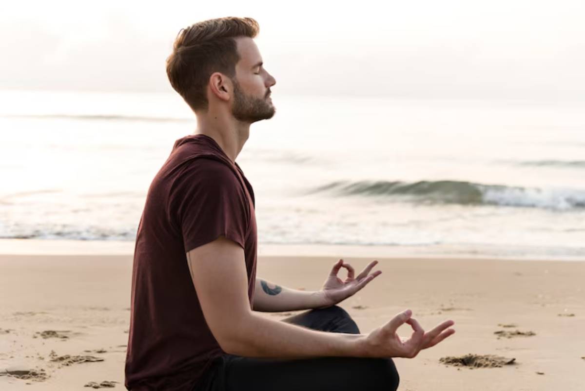 A person sits on the beach in a meditative pose, hands in a mudra, with gentle waves and a serene sky in the background.
