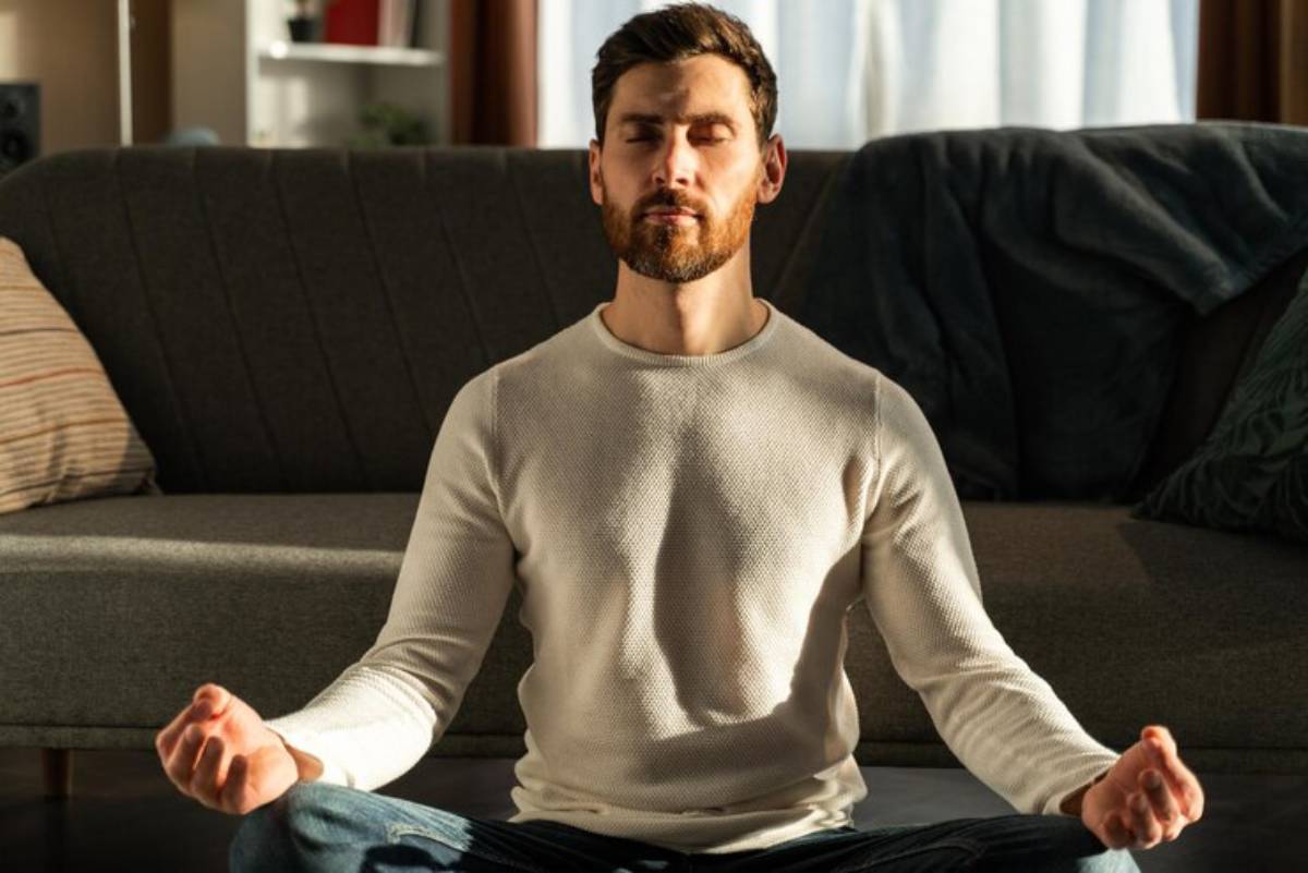 A person sits cross-legged on a sofa, meditating in a sunlit room with plants and soft furnishings in the background.