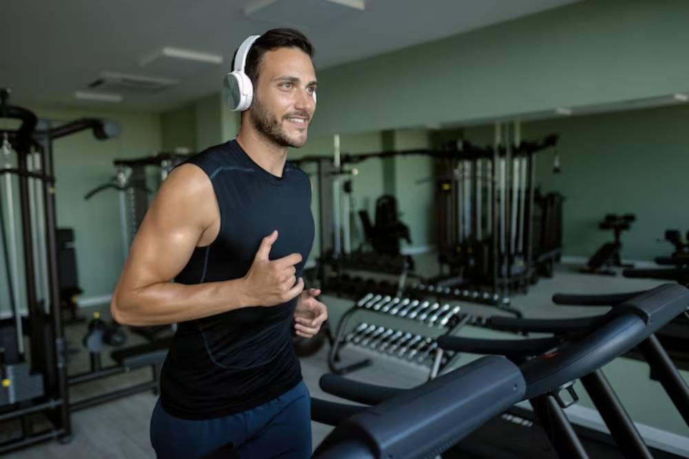 A person in a sleeveless shirt is jogging on a treadmill, wearing headphones in a modern gym surrounded by fitness equipment.