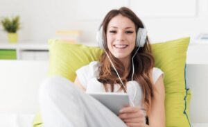 A person sitting on a couch with green pillows, wearing headphones, and holding a tablet. The room has a bright, minimalist design.