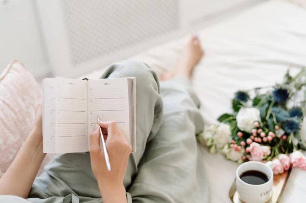 A person writes in a journal while sitting on a bed, surrounded by flowers and a cup of coffee.