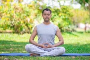 A person sits cross-legged on a yoga mat in an outdoor green setting, practicing meditation with hands in a mudra position.