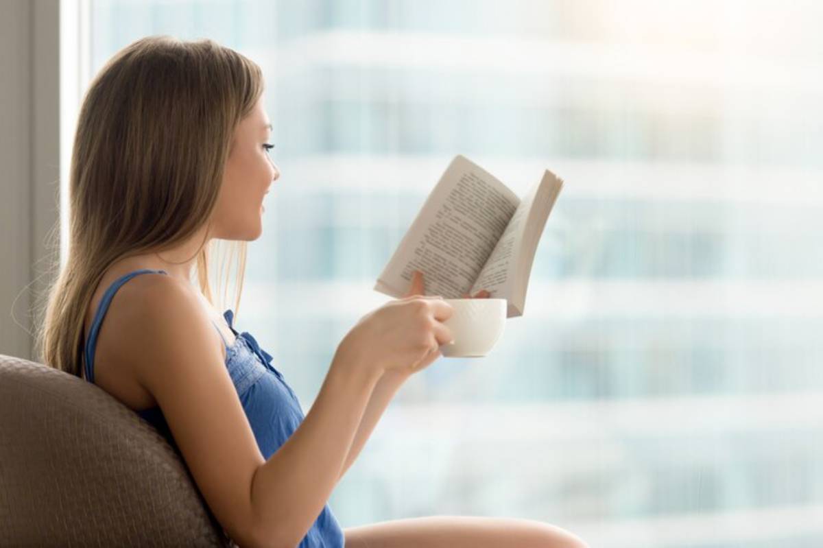 A woman in a blue tank top sits by a large window, sipping from a mug and reading a book, with a cityscape blurred in the background.