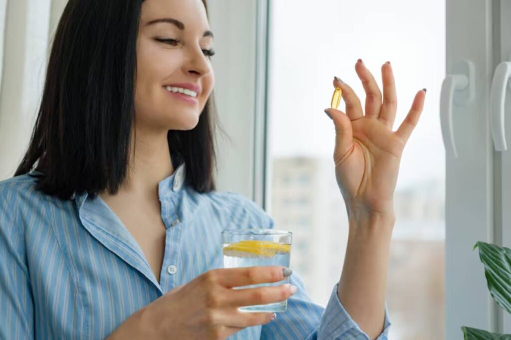 A person in a blue striped shirt holds a glass of lemon water in one hand and a yellow capsule in the other, standing by a window.