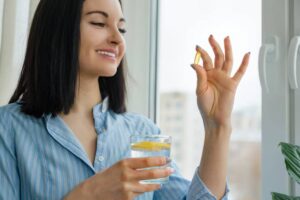 A person in a blue striped shirt holds a glass of lemon water in one hand and a yellow capsule in the other, standing by a window.