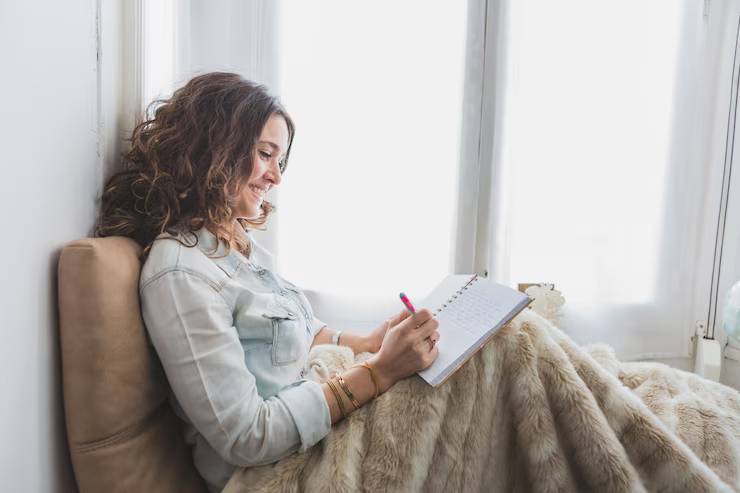 A woman with curly hair sits on a cozy chair, wrapped in a soft blanket, writing in a notebook by a bright window.