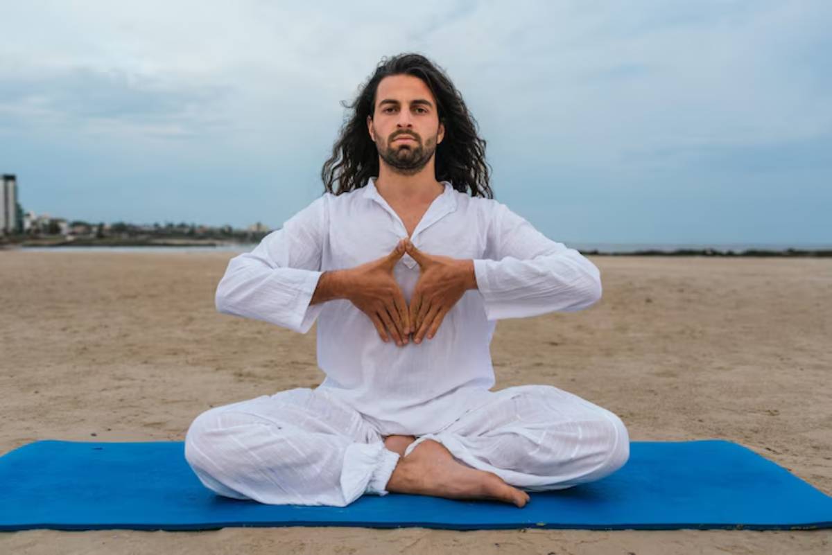A person in a white outfit sits cross-legged on a blue mat on the beach, practicing yoga with hands positioned in prayer.